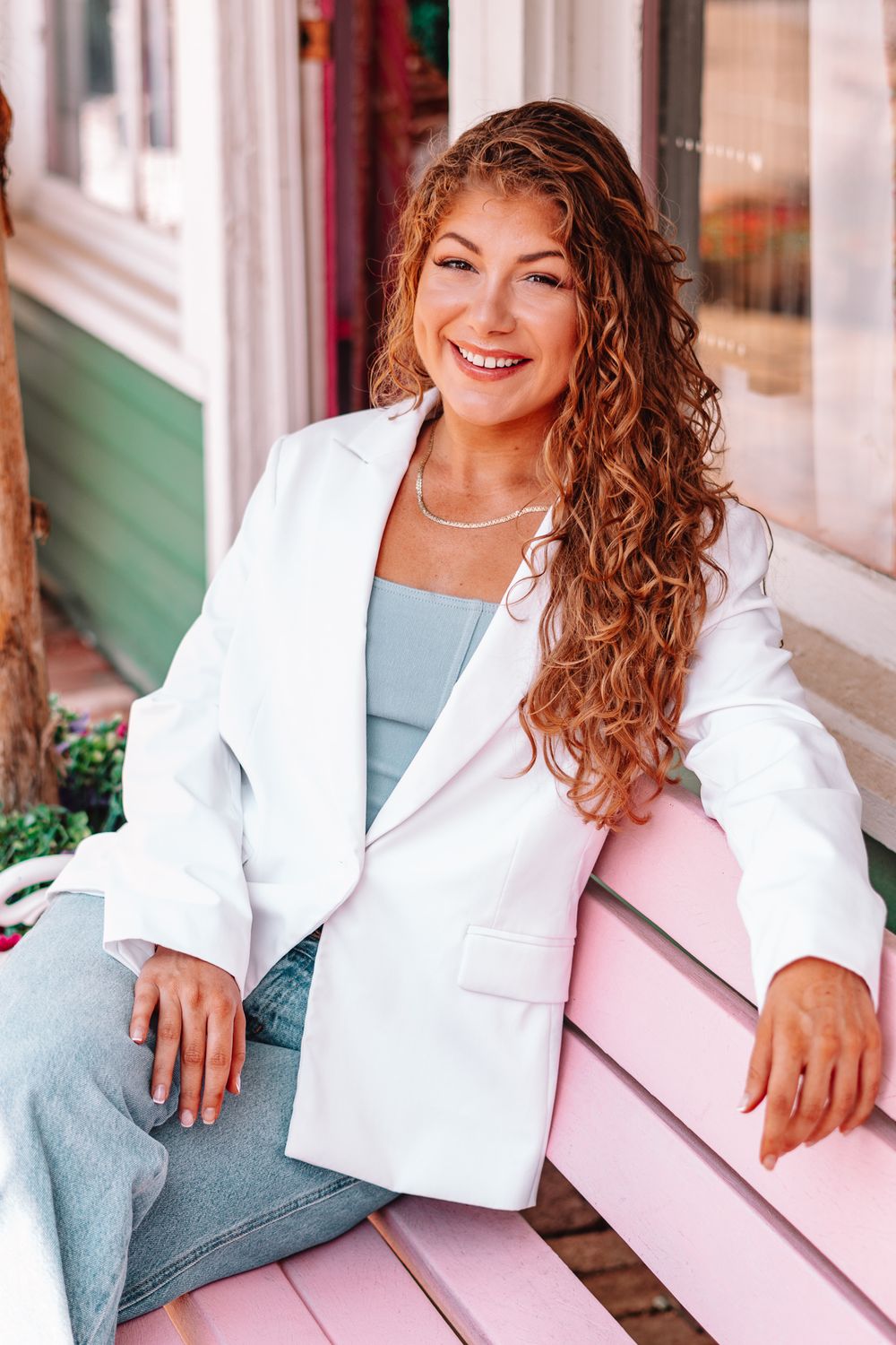 Woman in white blazer and blue jeans sitting casually on pink steps in an outdoor setting.