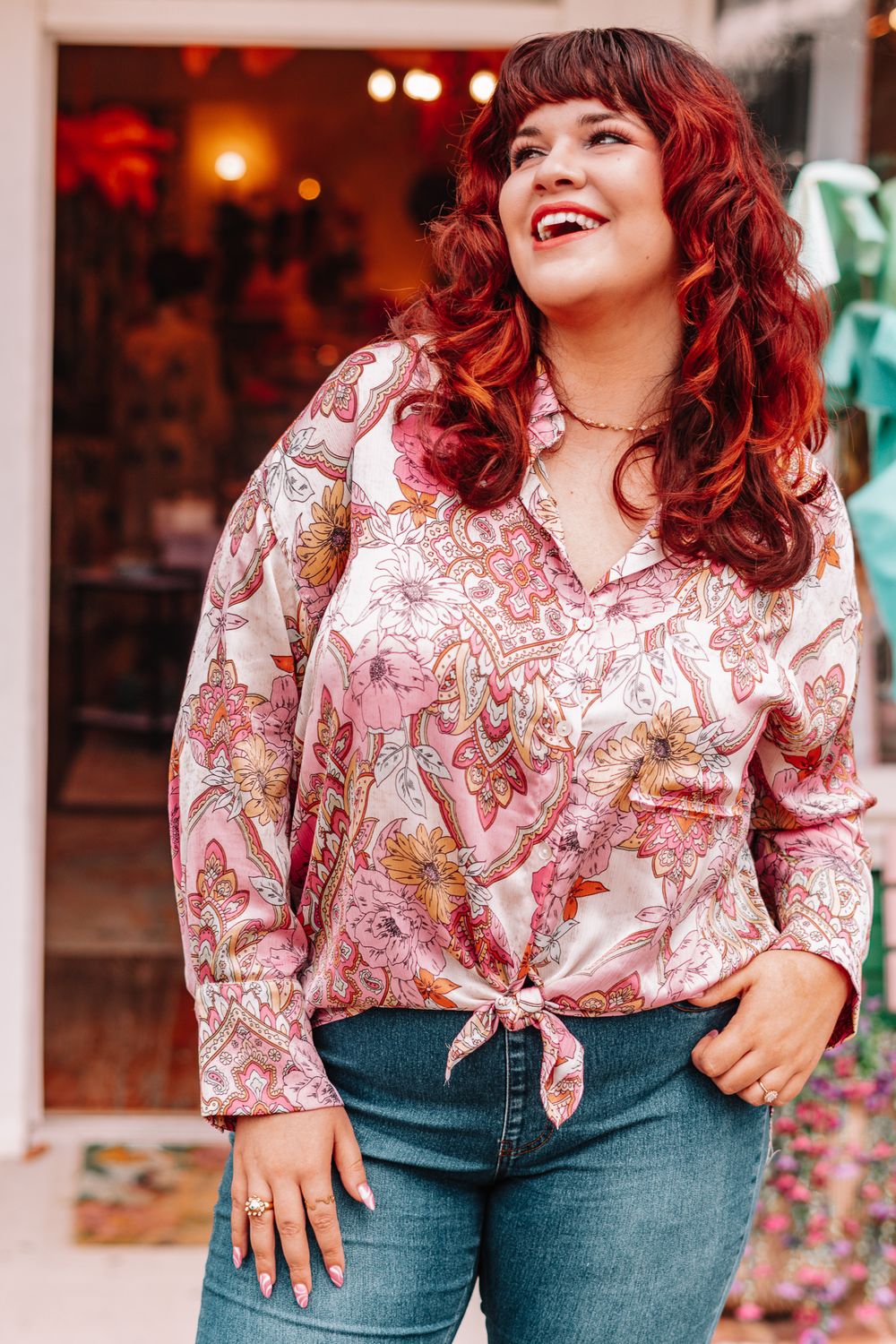 Woman in pink patterned top and jeans posing playfully against white porch railing.