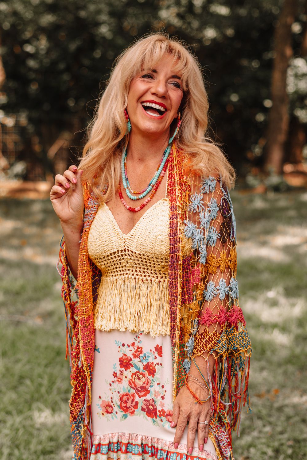 Woman in bohemian outfit with crochet vest and colorful shawl posing in garden setting.