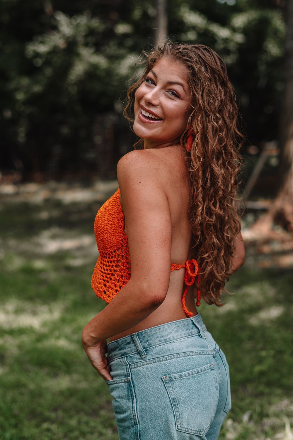 Woman in orange crop top and light denim poses playfully outdoors.
