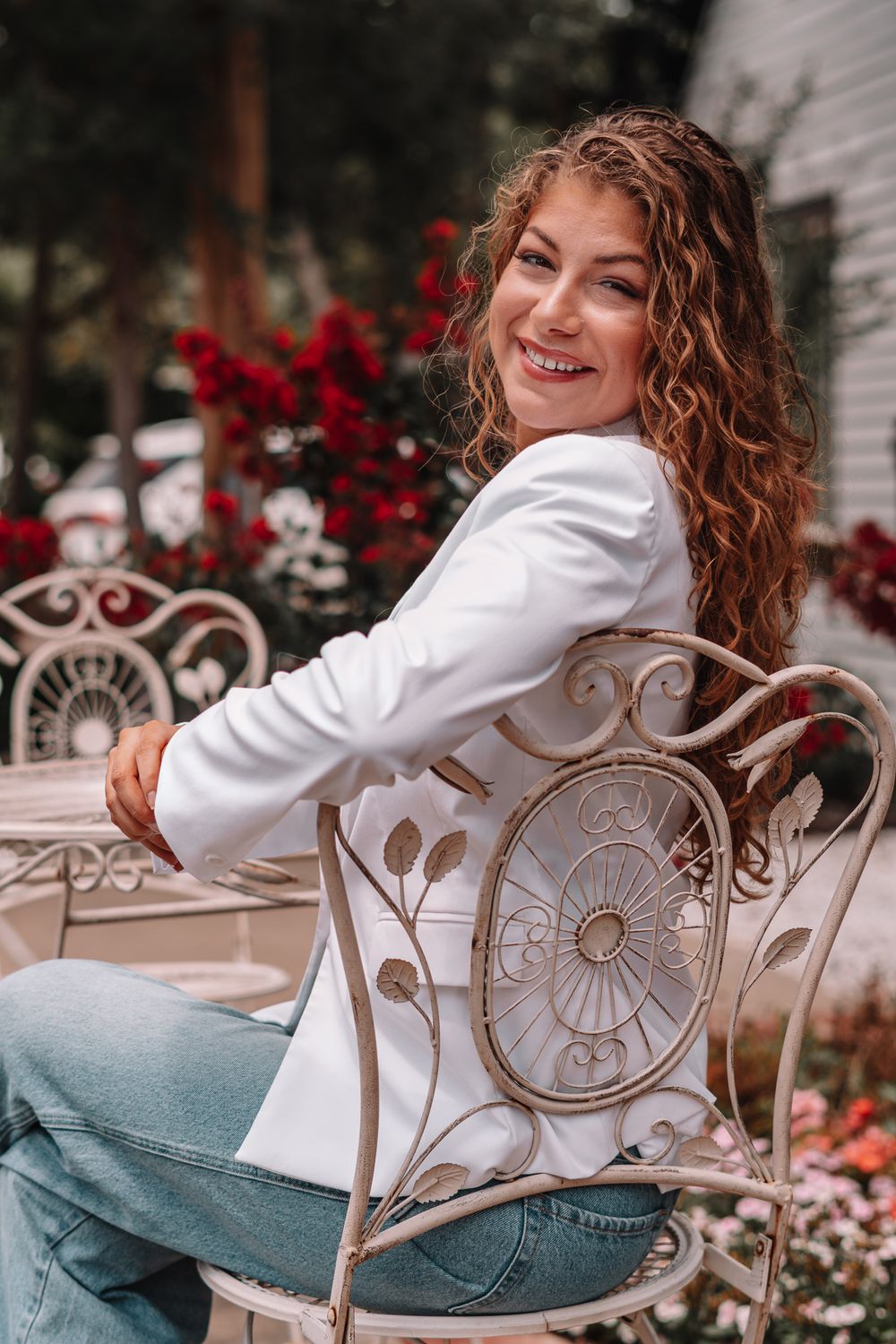 Woman relaxing in white blazer on decorative metal patio chair with red flowers in background.