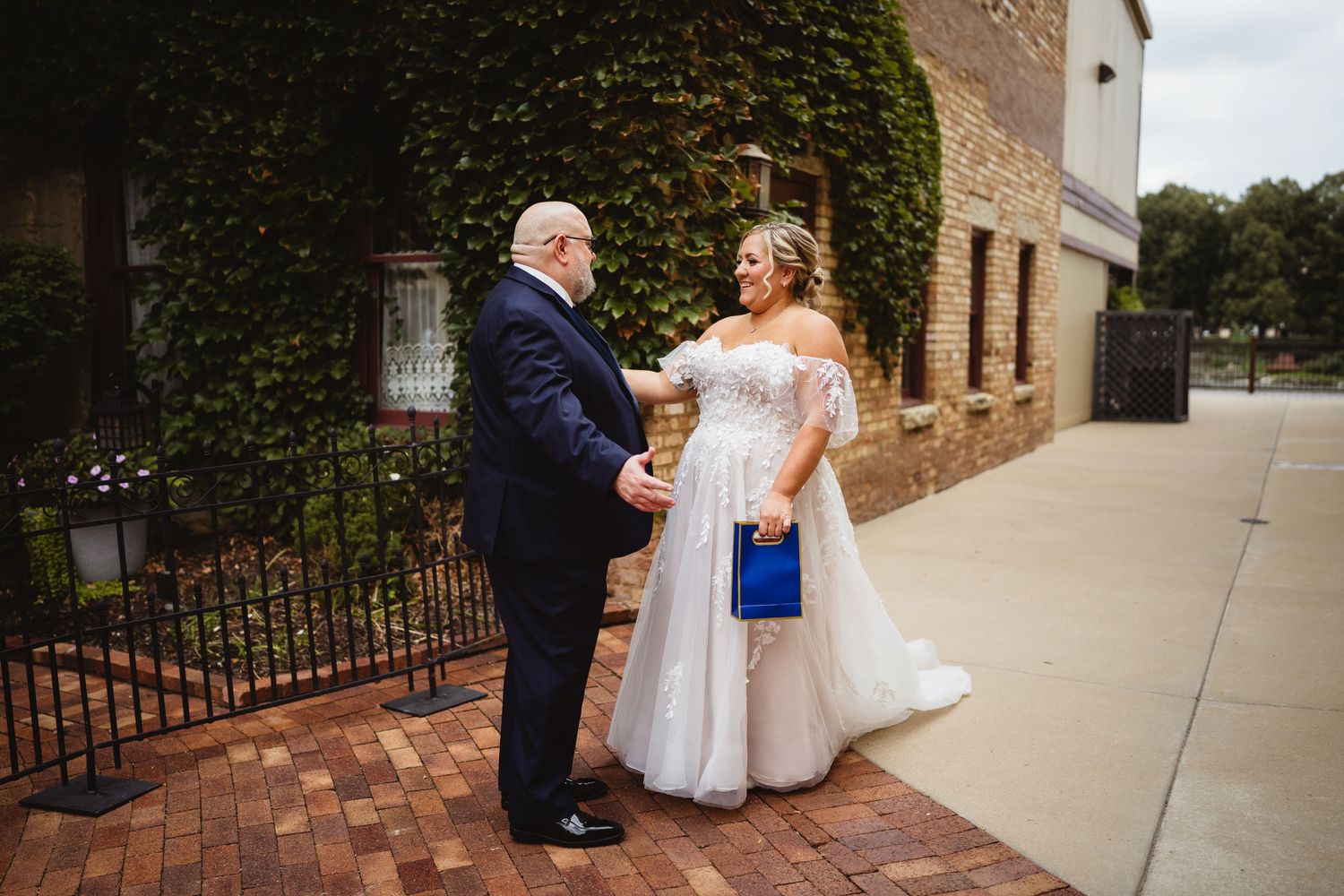 Two people sharing a moment together on a brick sidewalk, with one wearing a white wedding gown and holding a blue clutch.