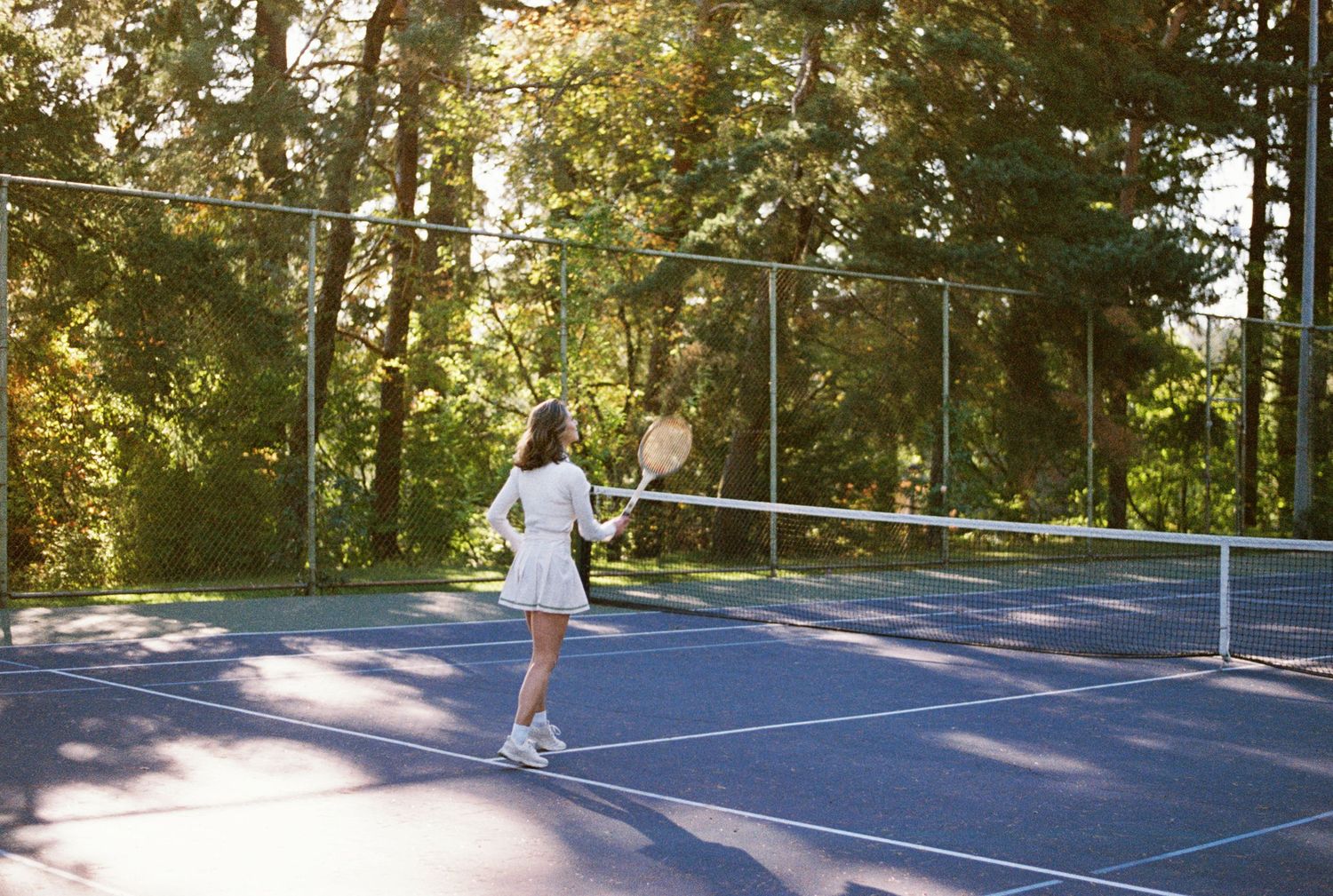 Several tennis players practicing serves on blue outdoor courts surrounded by tall green trees in sunlight.