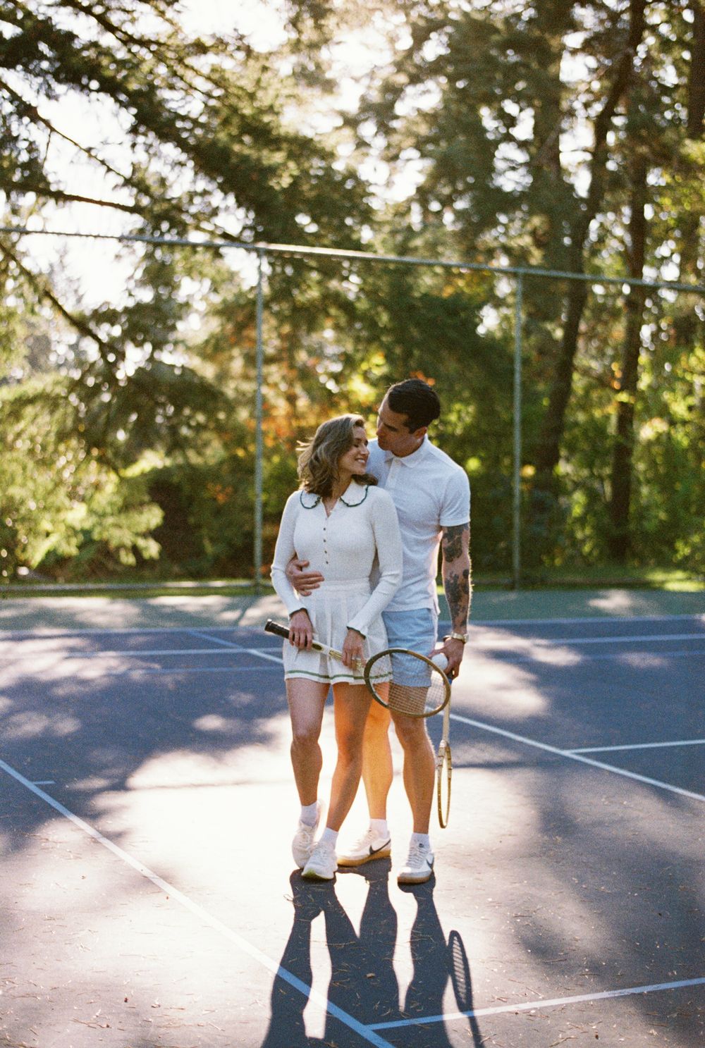 A romantic couple embraces on an outdoor tennis court during golden hour with long shadows on the ground.