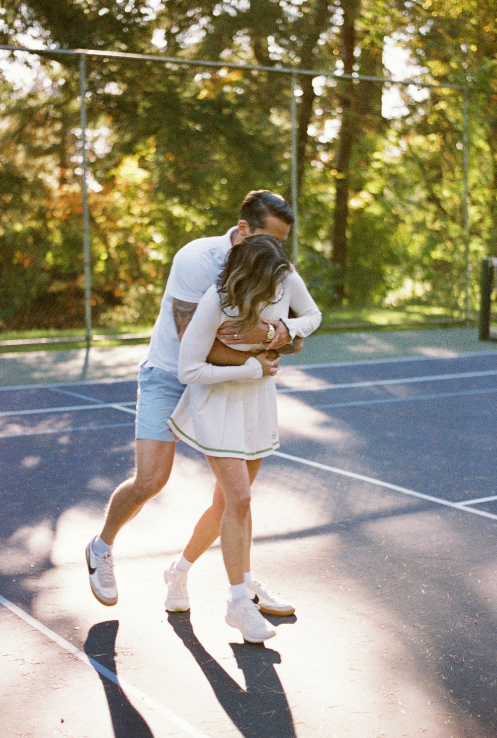 A playful couple in casual attire dancing and spinning together on an outdoor tennis court in golden sunlight.