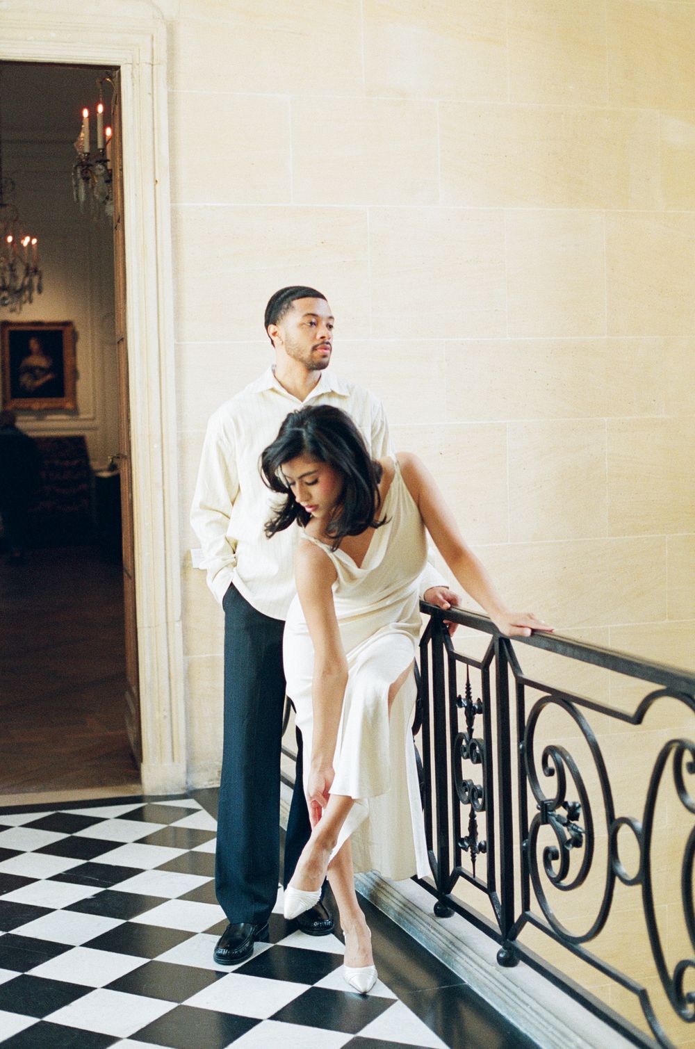 A couple in wedding attire pose playfully on a black and white checkered floor with an ornate iron railing.Paris film photographer.