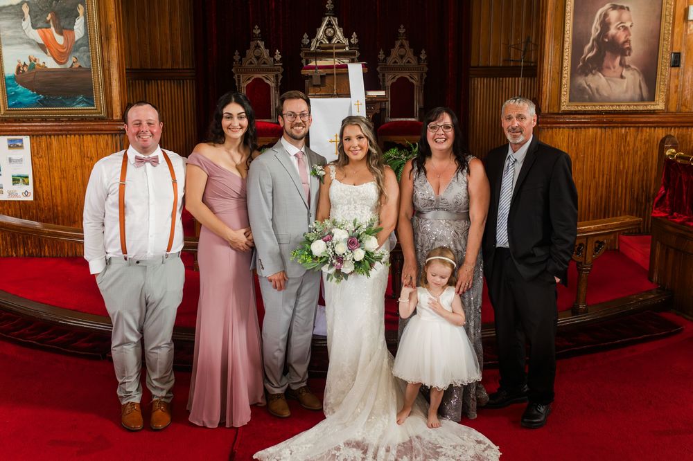 Wedding party members in pink dresses and formal attire pose together in church setting with decorative frames on wall.