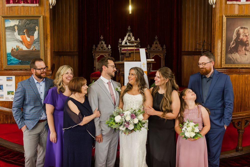 Wedding party members in formal attire gather inside a rustic wood-paneled venue for portrait photos.