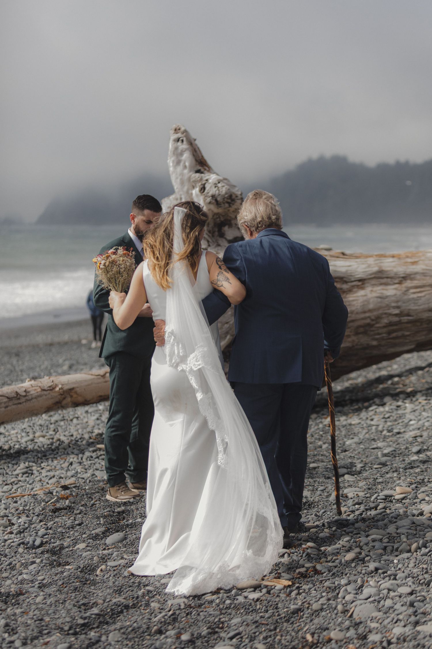 A wedding party stands along a foggy beach shoreline near a large driftwood log on a misty day.