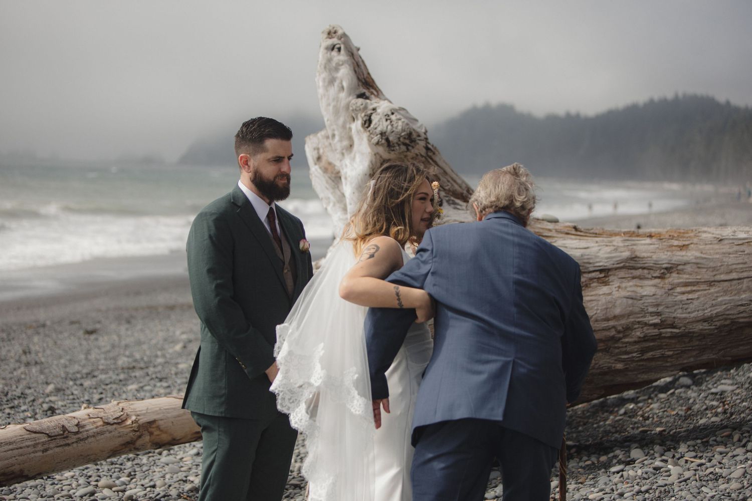 A scenic beach wedding ceremony takes place on a misty Pacific Northwest shoreline with driftwood and mountains in background.