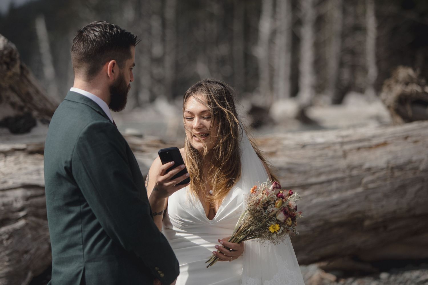 A couple in wedding attire looks at a phone together while standing in a dramatic forest landscape.
