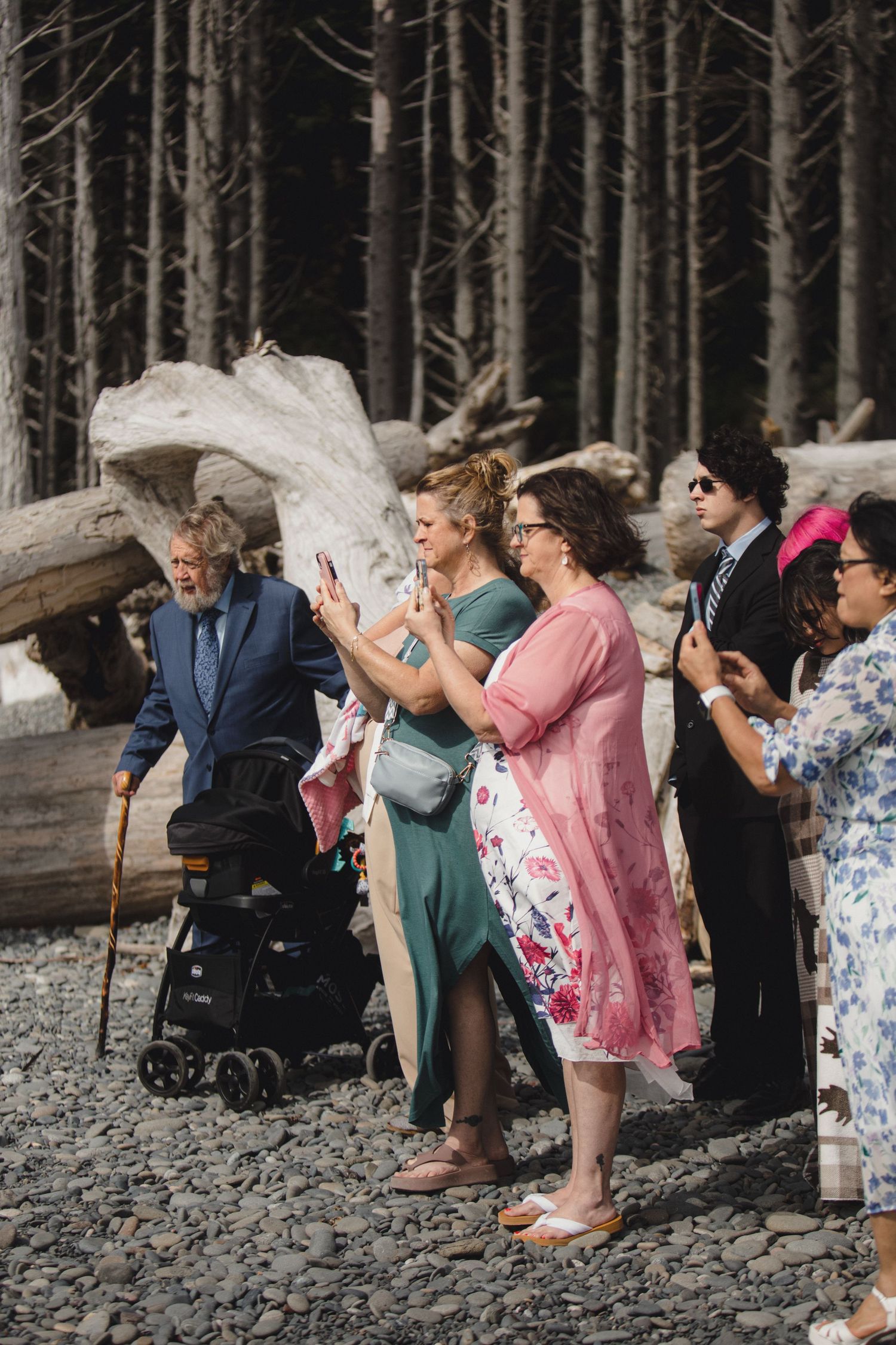 Wedding party poses dramatically leaning back in unison on rocky beach with forest backdrop.