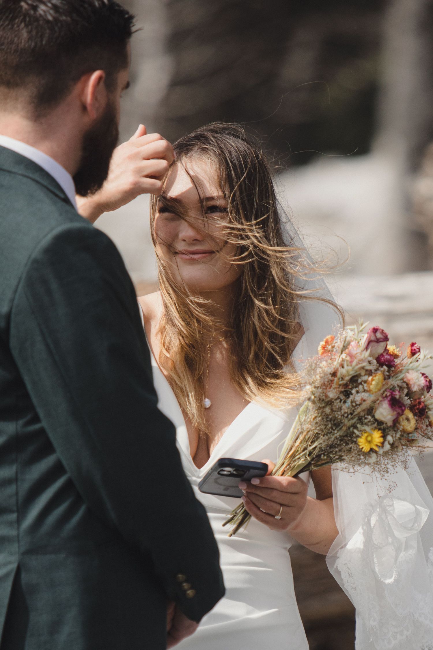 A tender moment between newlyweds outdoors as wind blows through her hair while she holds a dried flower bouquet.