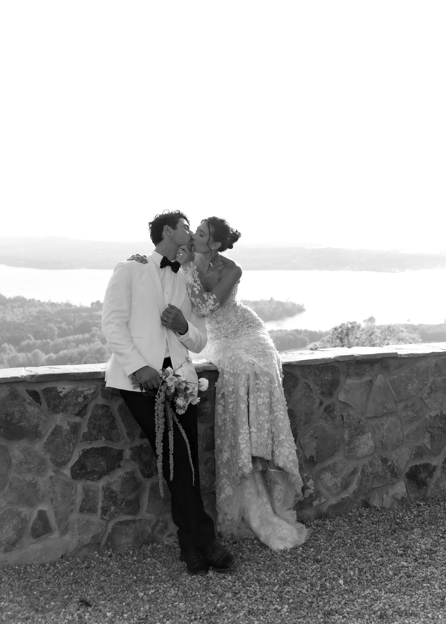 Black and white photo of a bride and groom sharing a kiss on a stone terrace with a scenic lake view behind them.