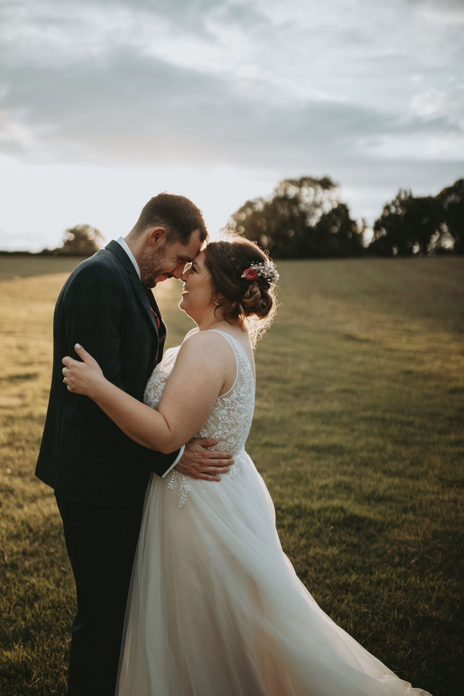 Romantic newlywed couple embraces in a sunset field wearing traditional wedding attire and sharing an intimate moment.