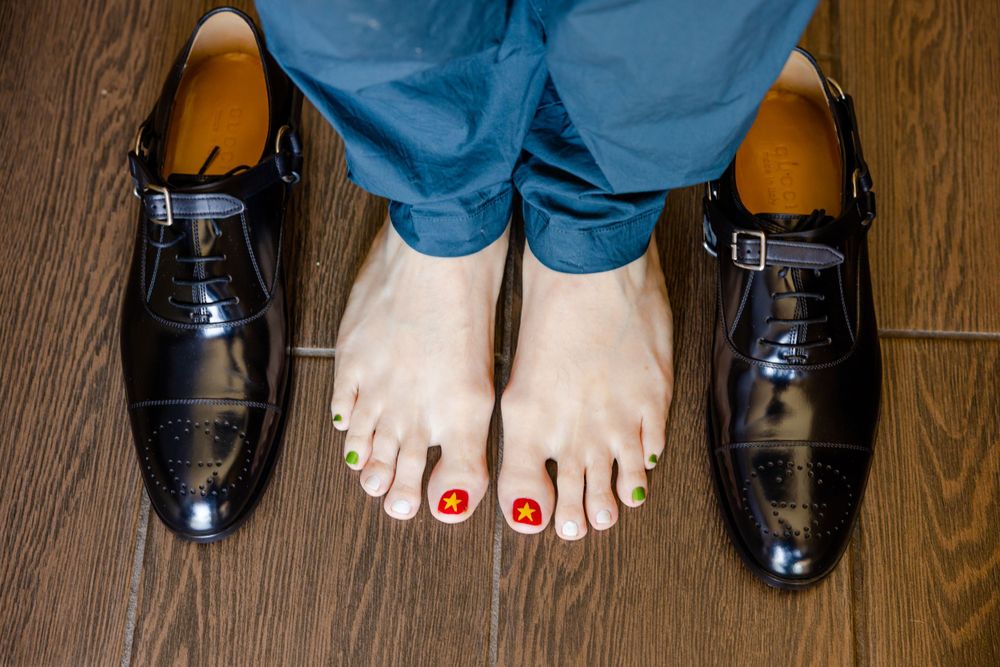 A pair of formal black dress shoes placed next to bare feet with red nail polish.