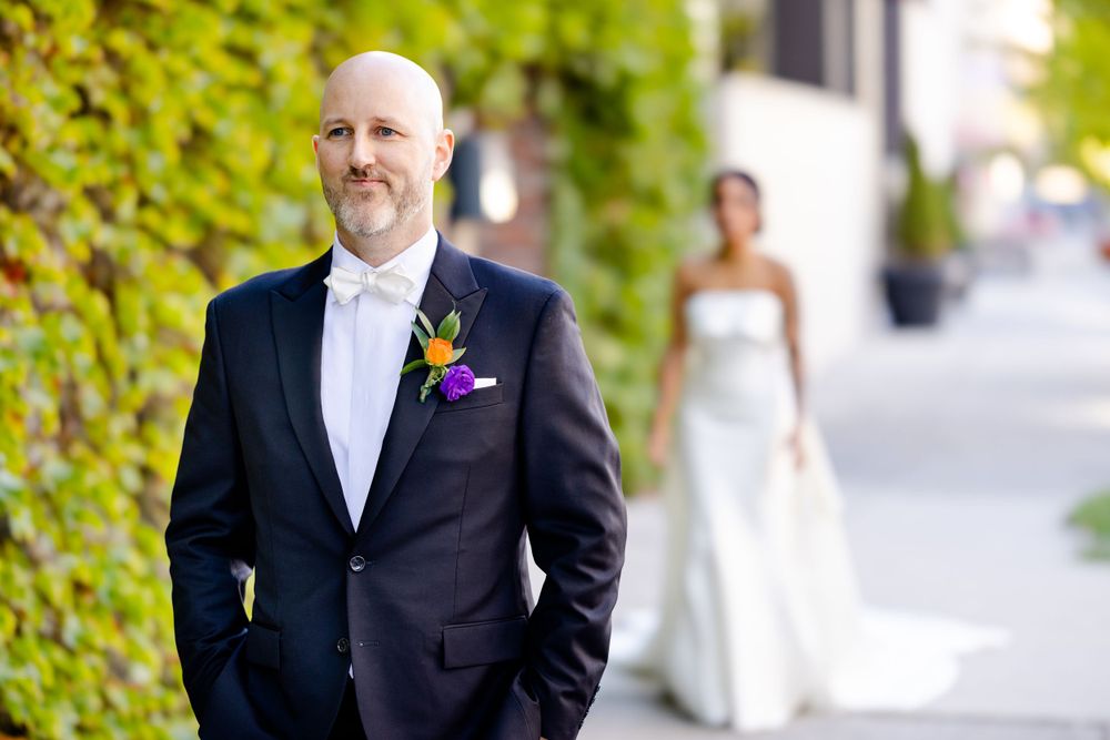 A groom in black tuxedo with white bowtie stands against ivy-covered wall with bride visible in background.