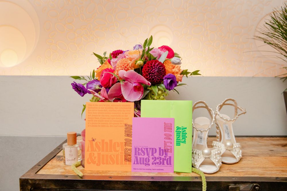 Colorful wedding bouquet with pink and purple flowers next to invitation cards and bridal shoes.