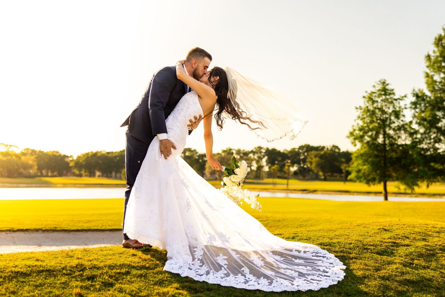 A wedding couple shares an intimate kiss in a golden sunlit field at sunset.