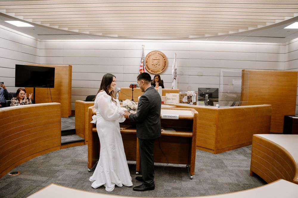A couple in wedding attire exchange vows in a simple courthouse setting with wooden paneled walls.