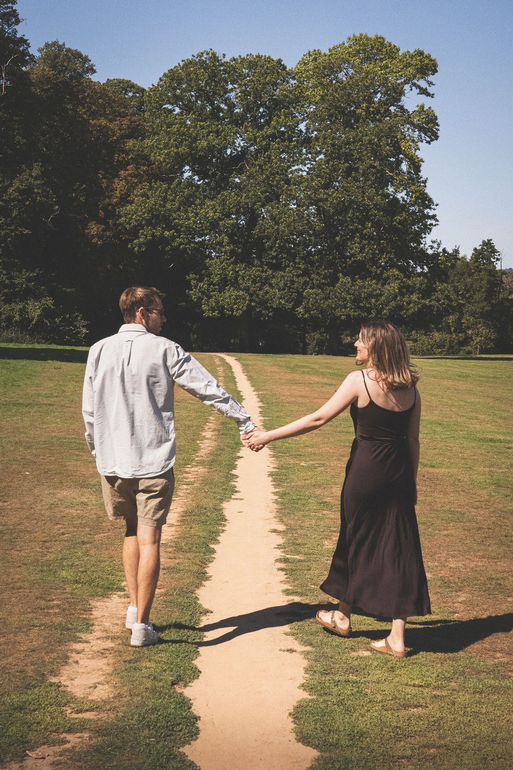 A couple holding hands walks along a dirt path through a sunny park with tall trees in the background.