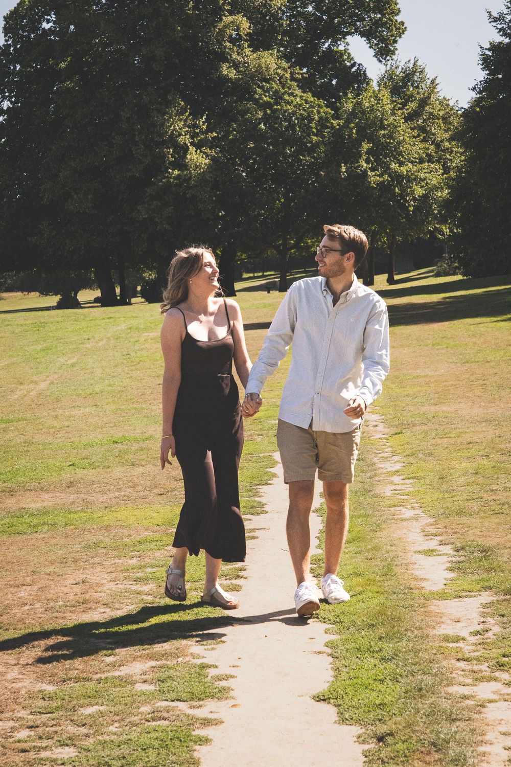 Two people stroll together on a walking path through a grassy park on a bright summer day.
