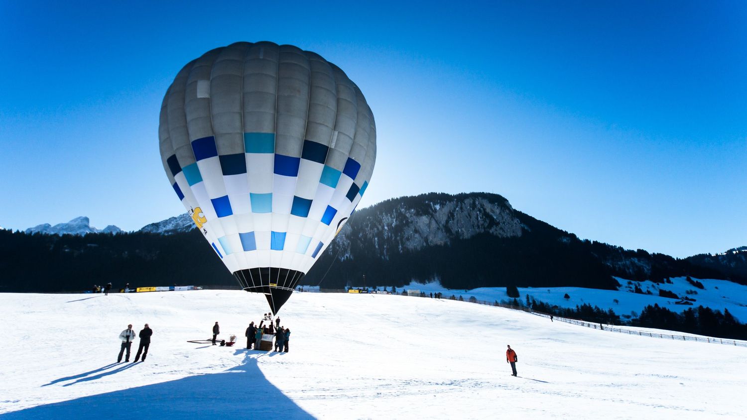 Hot air balloon floating above snowy mountains while skiers cross country ski on pristine white slopes below.