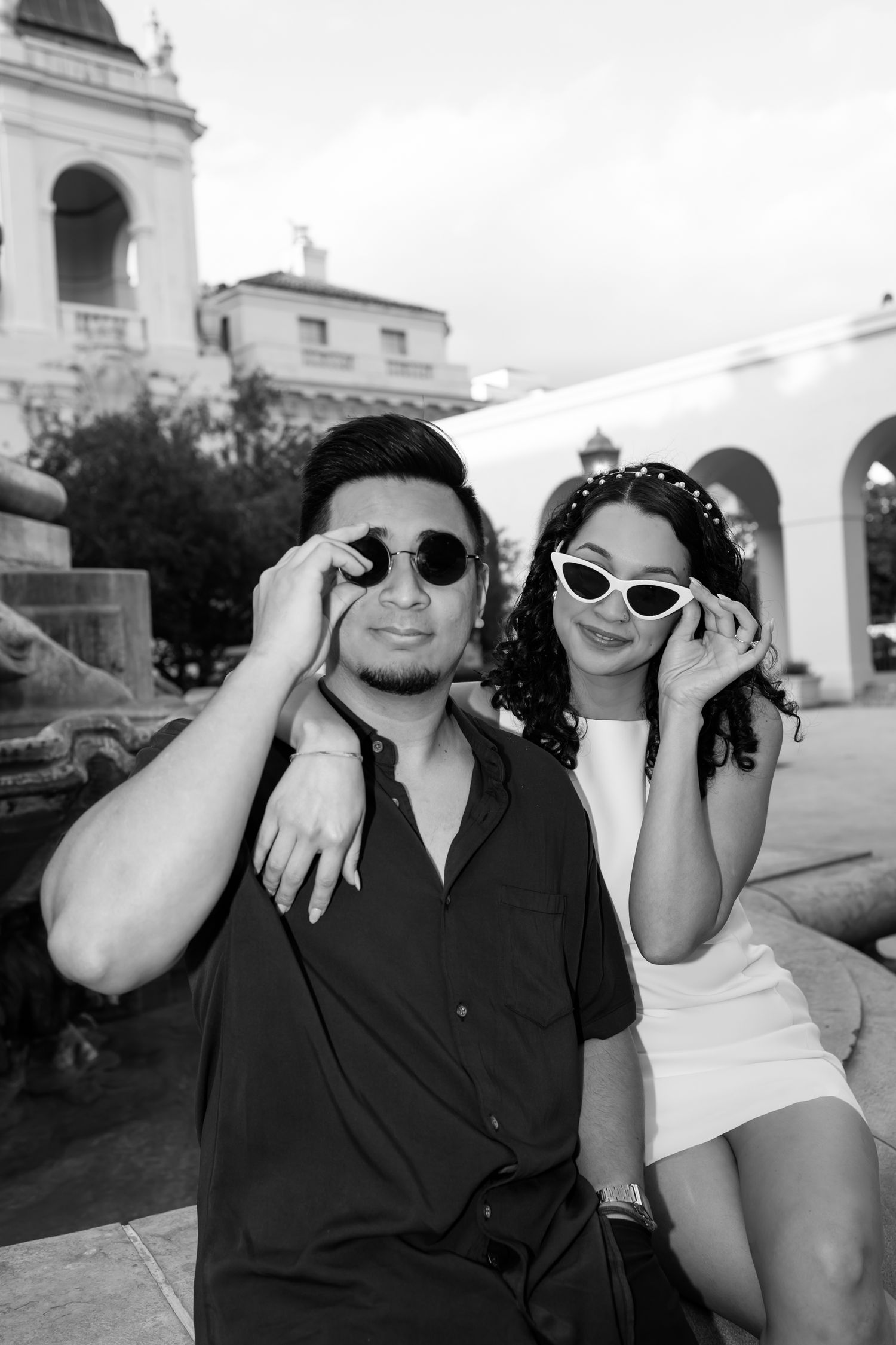 A stylish couple in sunglasses pose together outdoors in black and white, with architectural details visible in background.