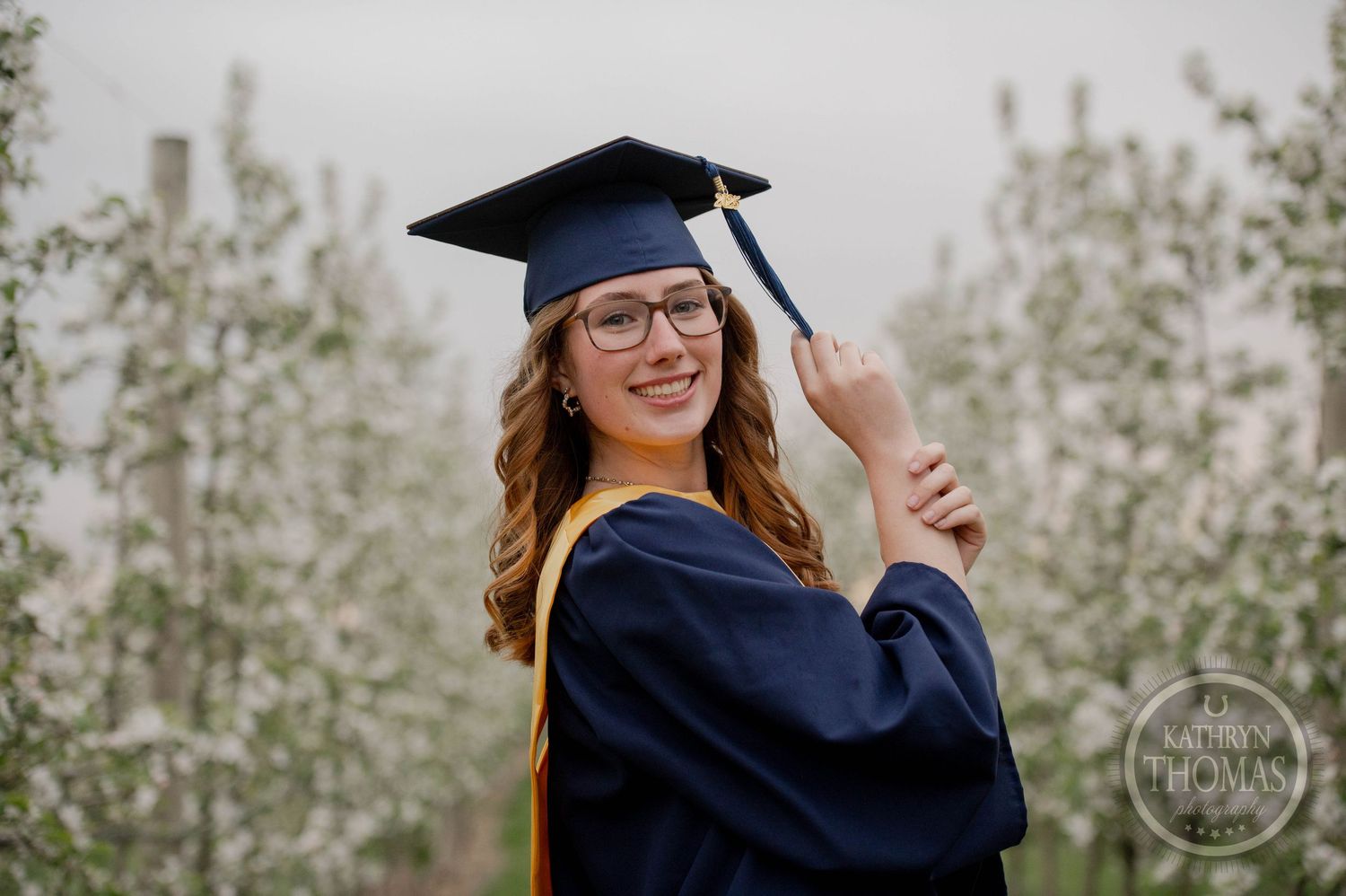 Graduate in navy cap and gown adjusts their mortarboard while smiling outdoors against a natural background.
