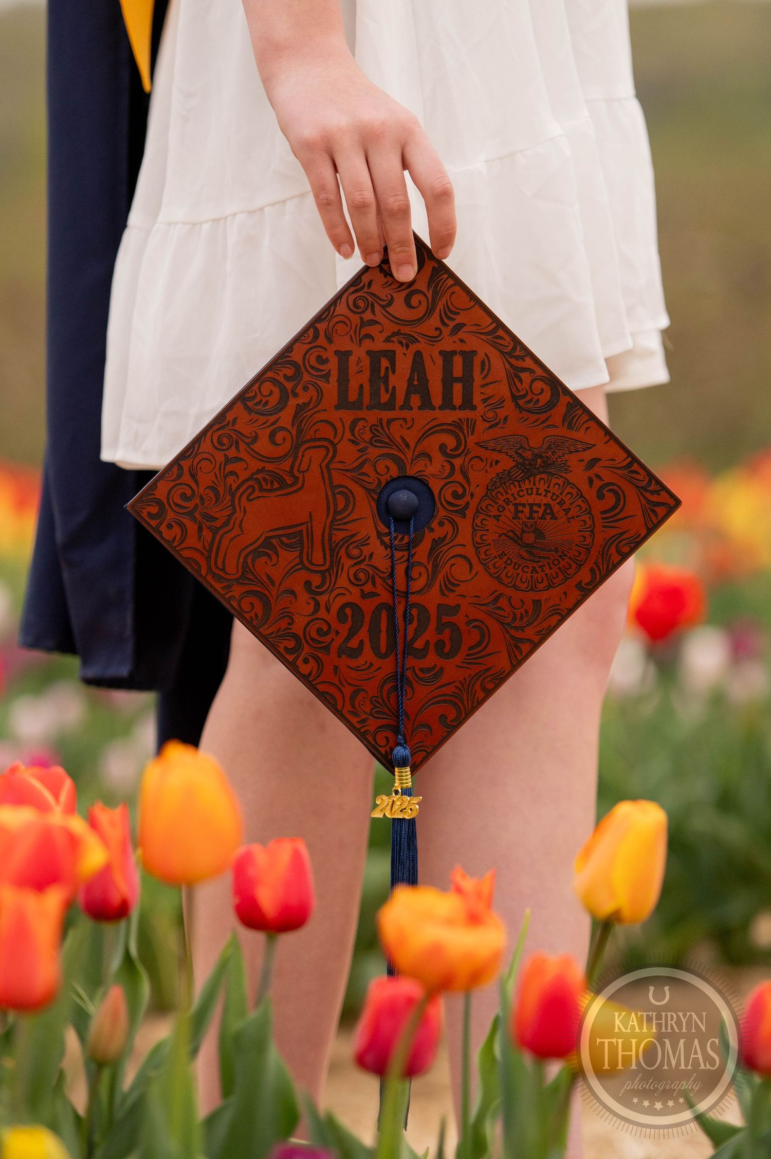 A decorated graduation cap with brown leather design sits among vibrant orange and red tulips in a garden setting.