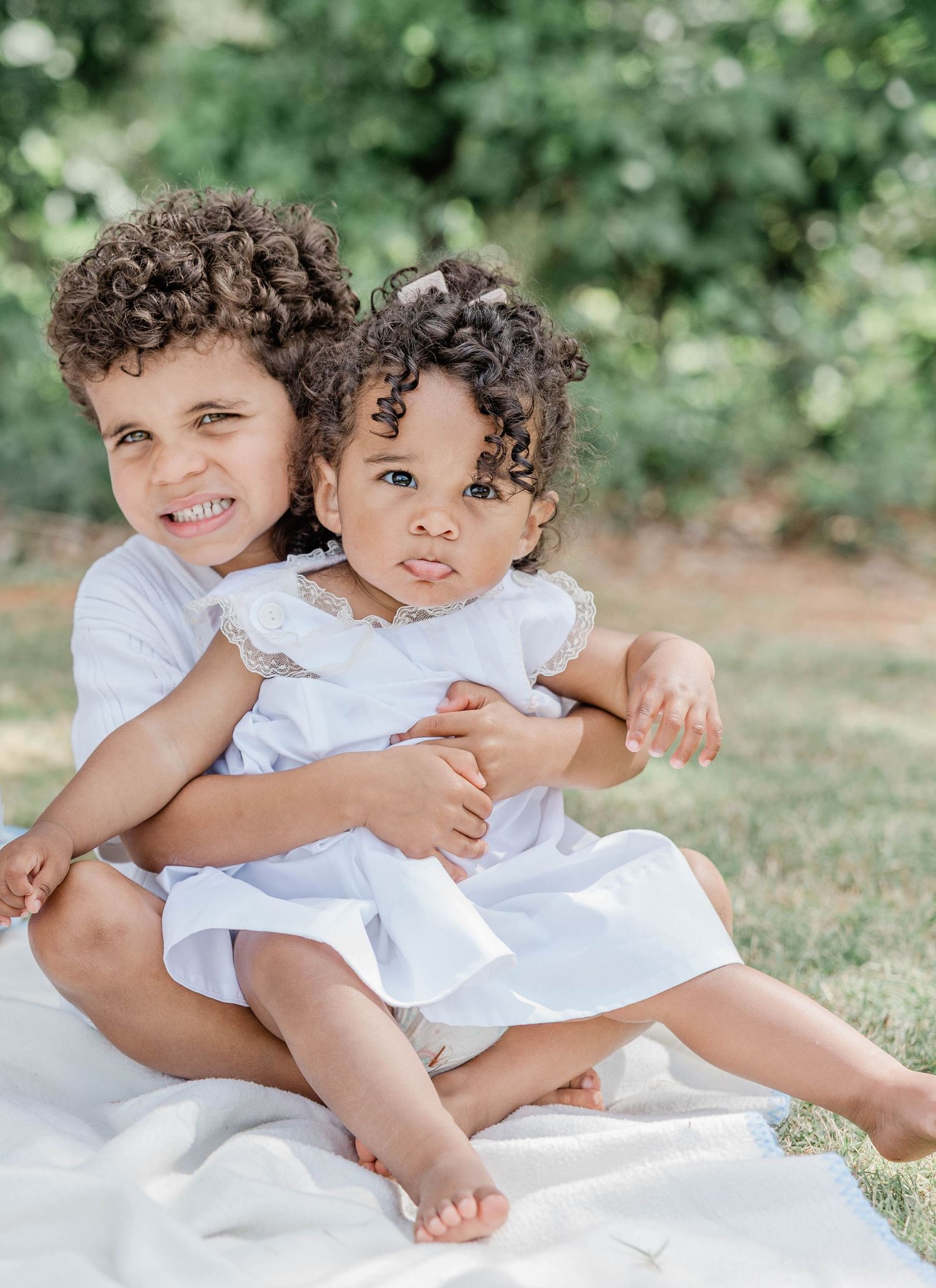 Two children in white outfits sitting on blanket outdoors.