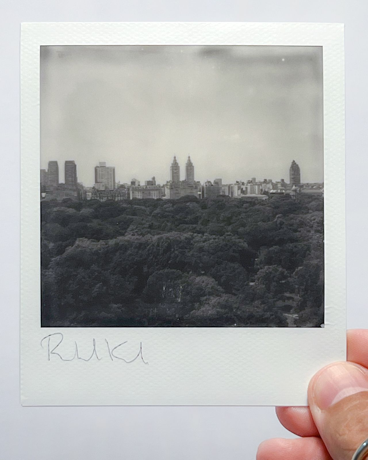 Black and white polaroid of Manhattan skyline viewed across Central Park's tree canopy.