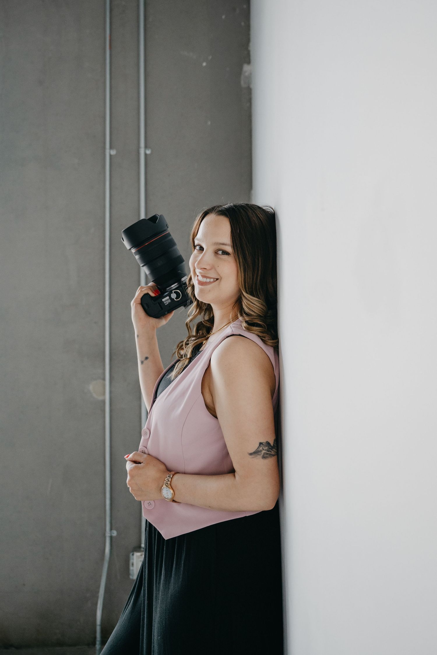 Professional photographer in pink sleeveless top holds DSLR camera while posing against grey and white wall.
