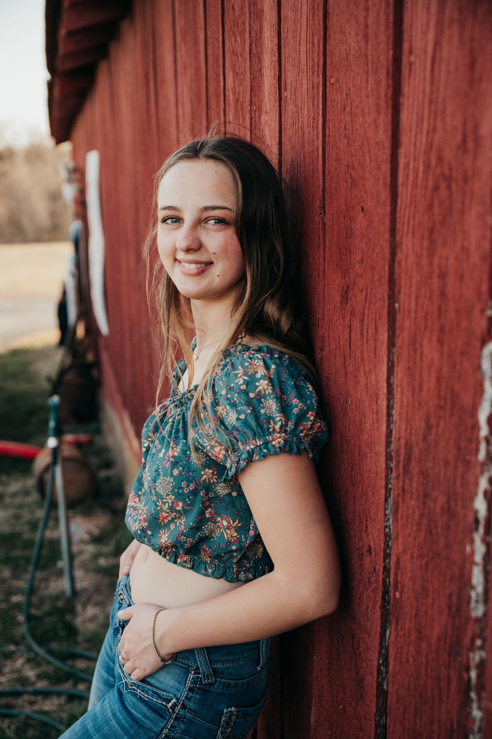 Young person in floral blue top and jeans poses against rustic red barn wall in outdoor portrait.