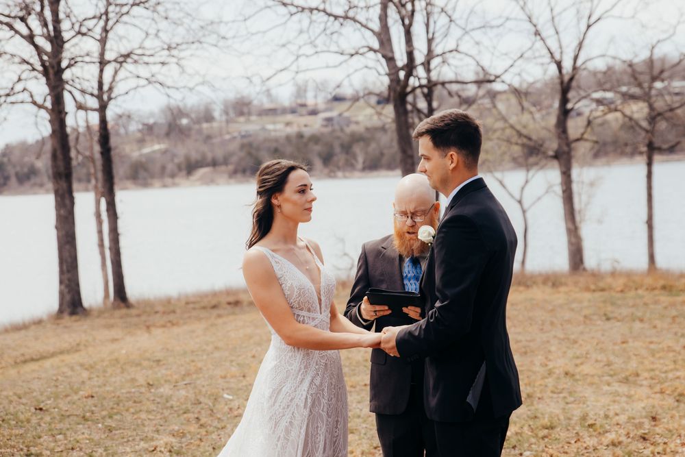 A wedding ceremony takes place outdoors by a lake in autumn with bare trees in the background.