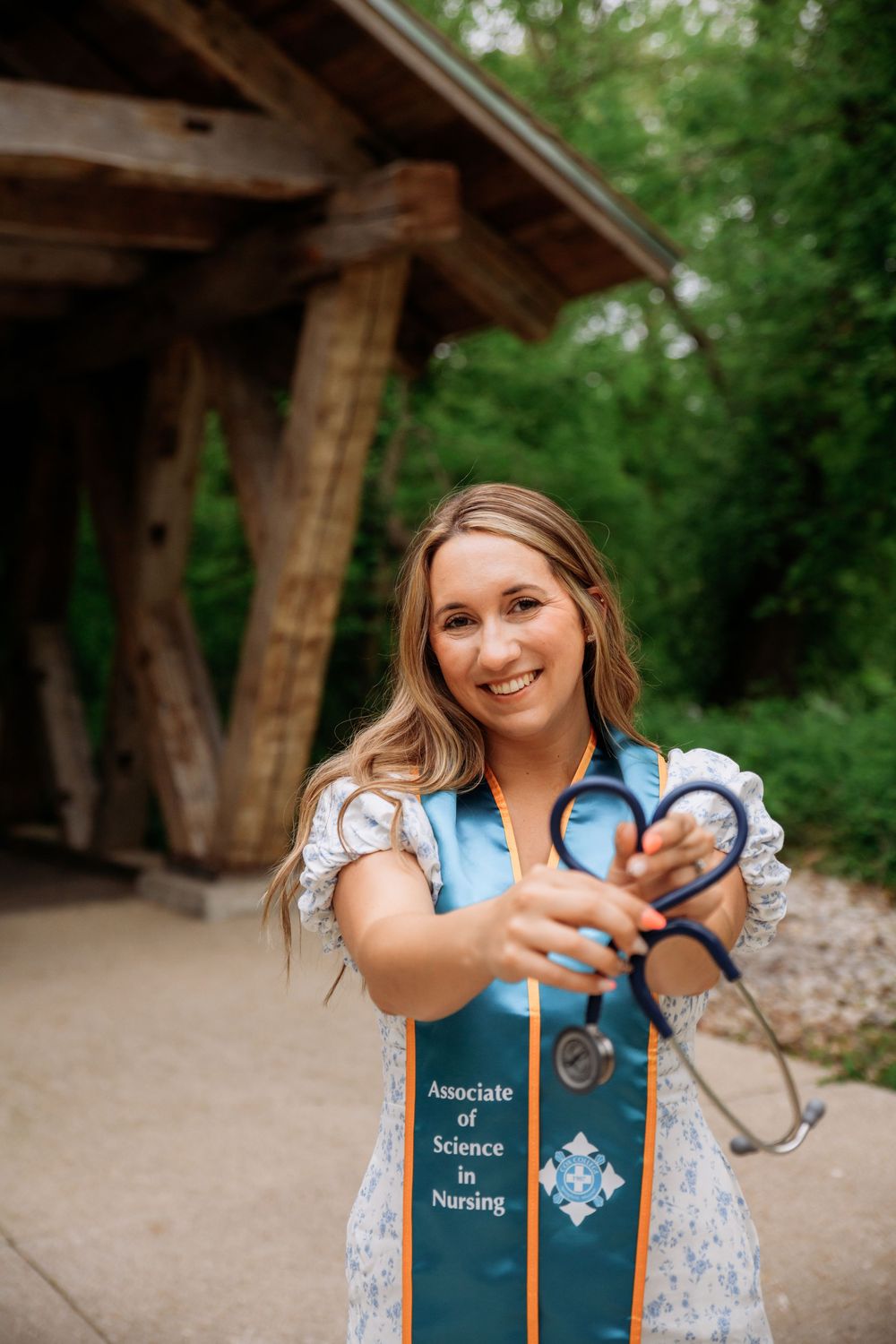 Graduate in nursing wearing a graduation stole and holding a stethoscope stands smiling under a wooden structure outdoors.