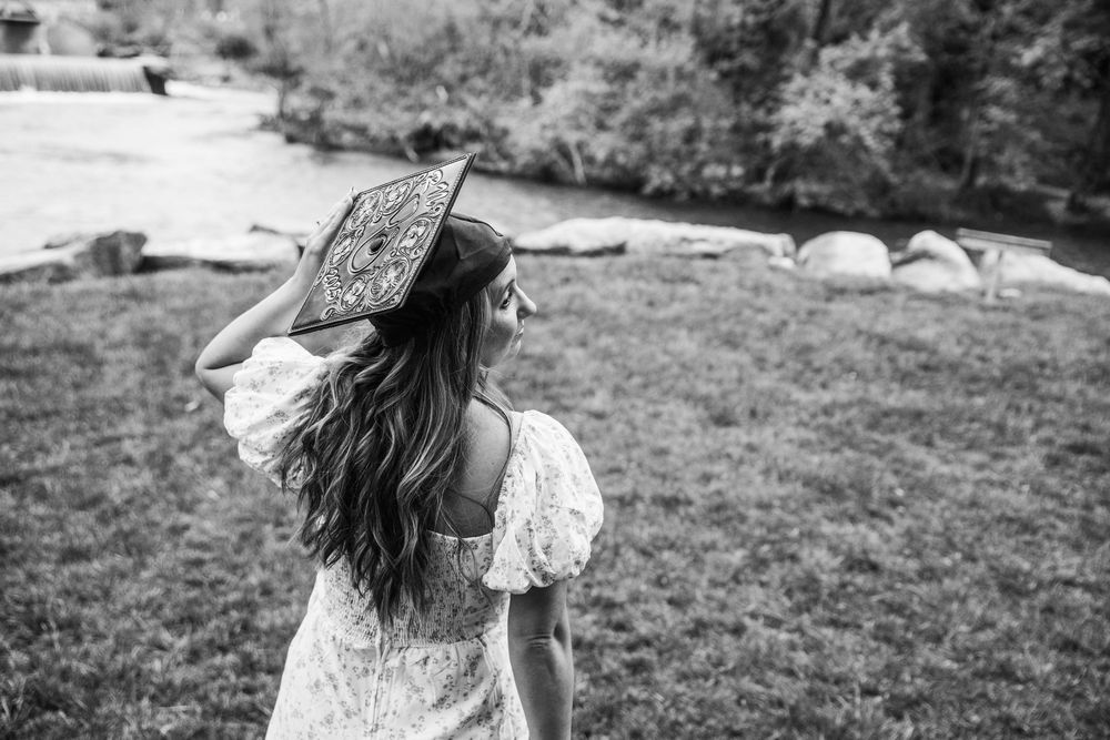 Person in a flowy white dress and wide-brimmed hat stands in field with long hair flowing in black and white image.