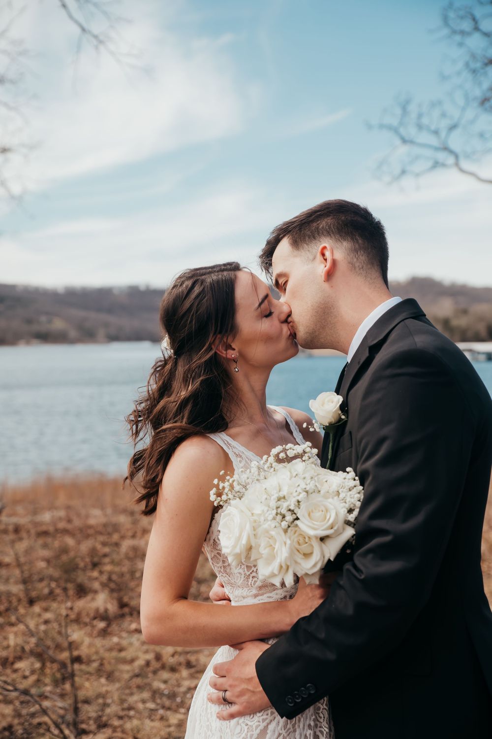 Newlyweds share romantic kiss by lakeside during autumn wedding with bride holding white rose bouquet.