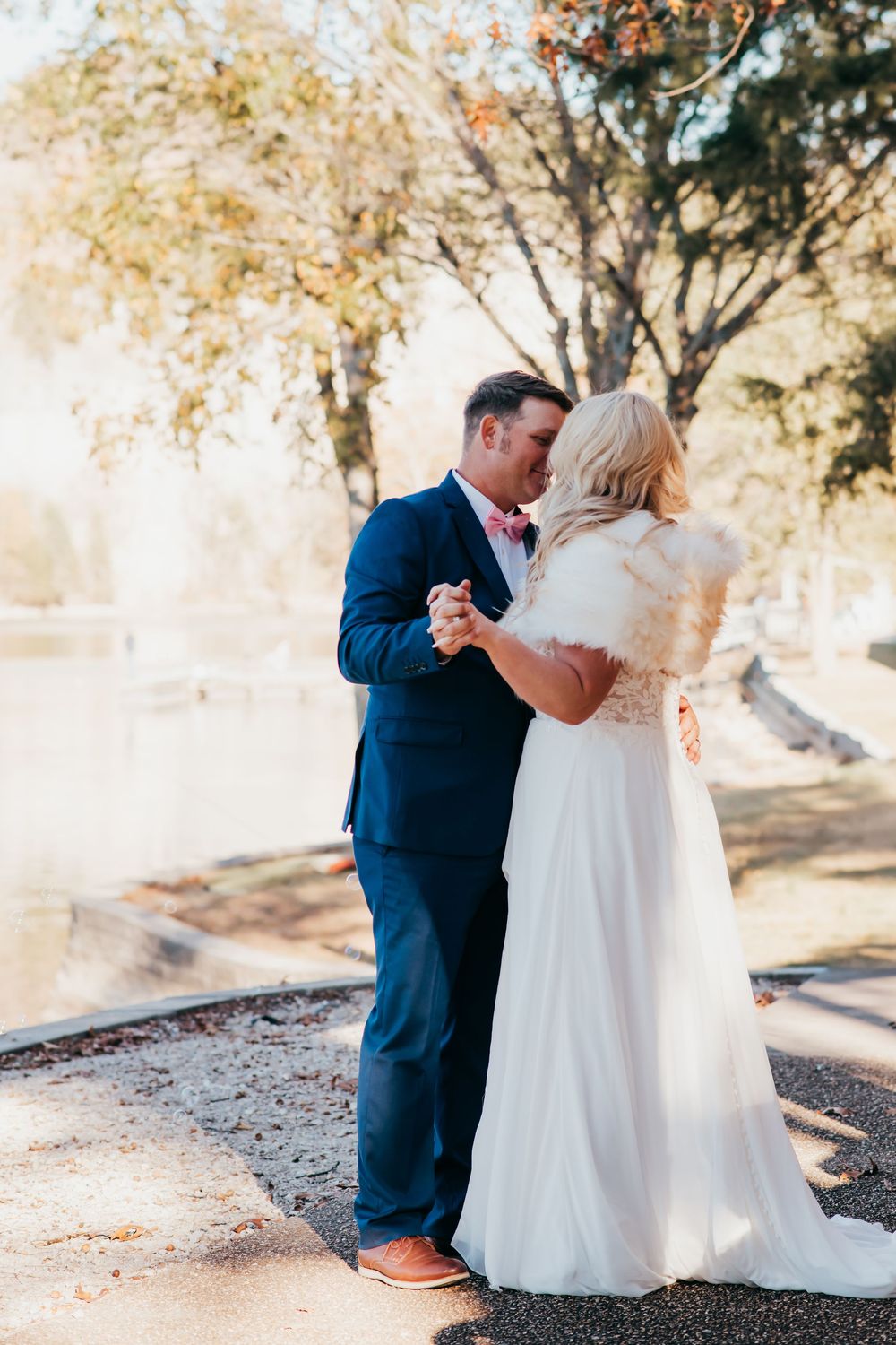 A couple in wedding attire dance together outdoors on a sunlit path surrounded by autumn trees.