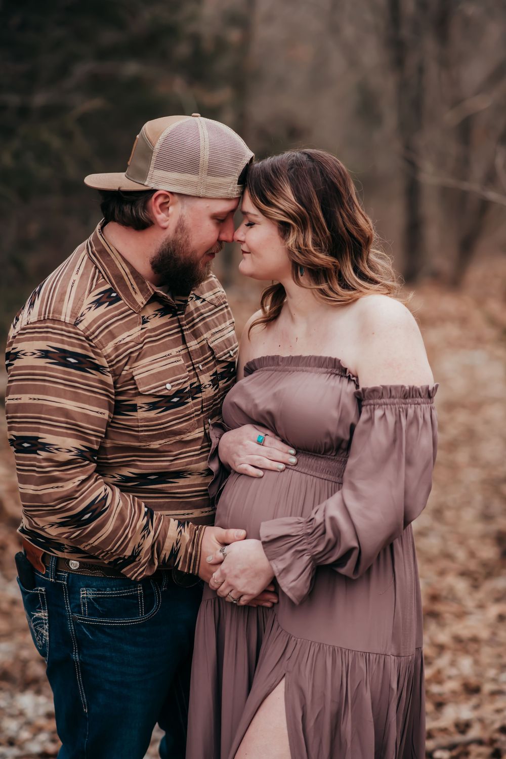 Intimate autumn maternity photo shows couple embracing in brown tones among fallen leaves in forest setting.