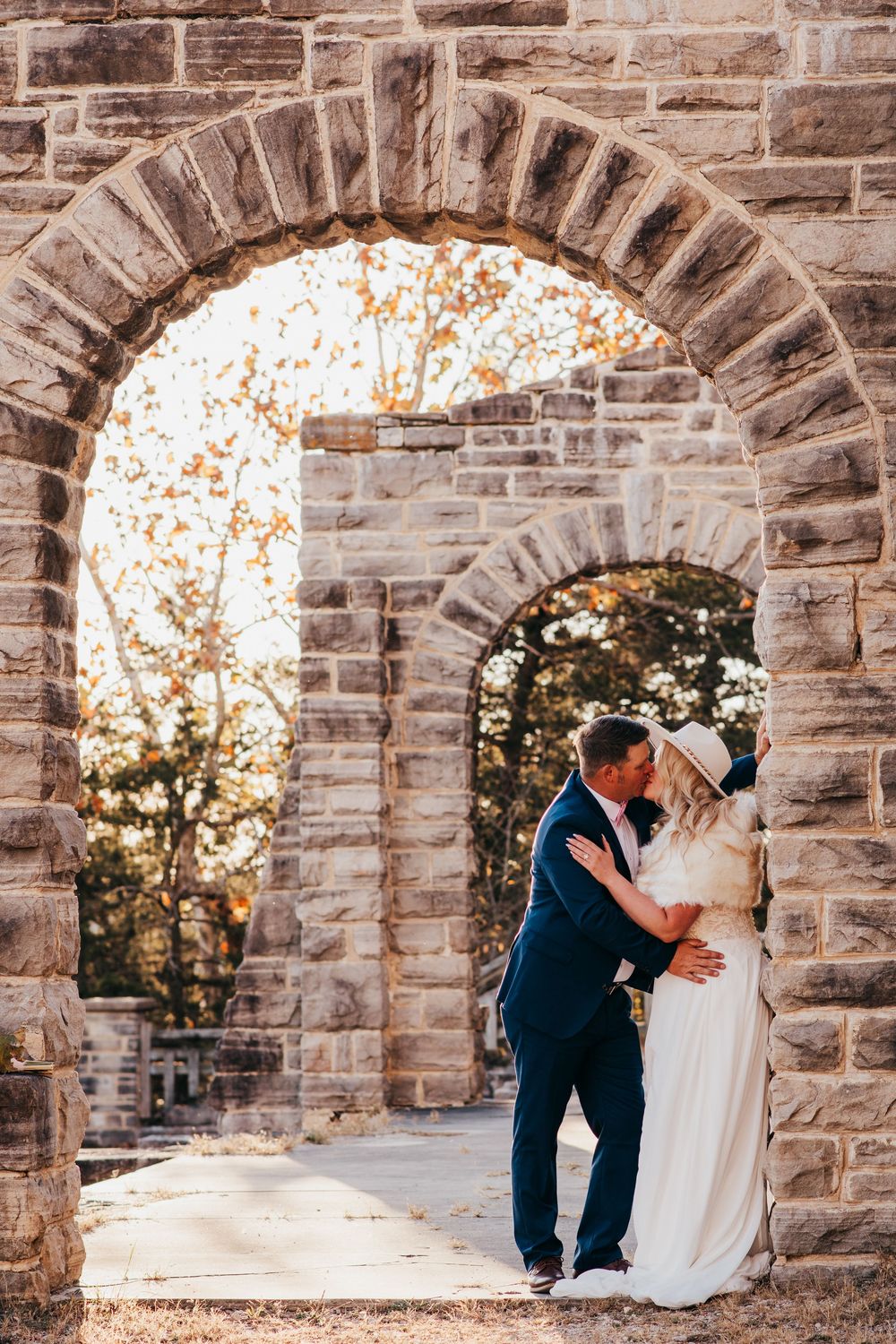 Romantic moment captured under stone archways with autumn foliage glowing in golden sunlight.