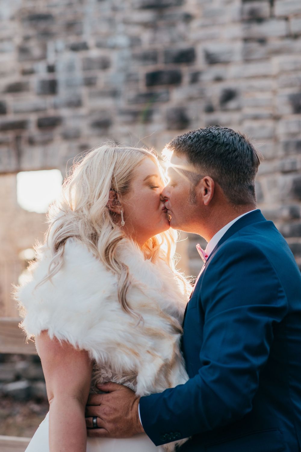 A romantic sunset wedding photo of a bride in white with a fur wrap kissing their partner in a navy suit against a stone wall.