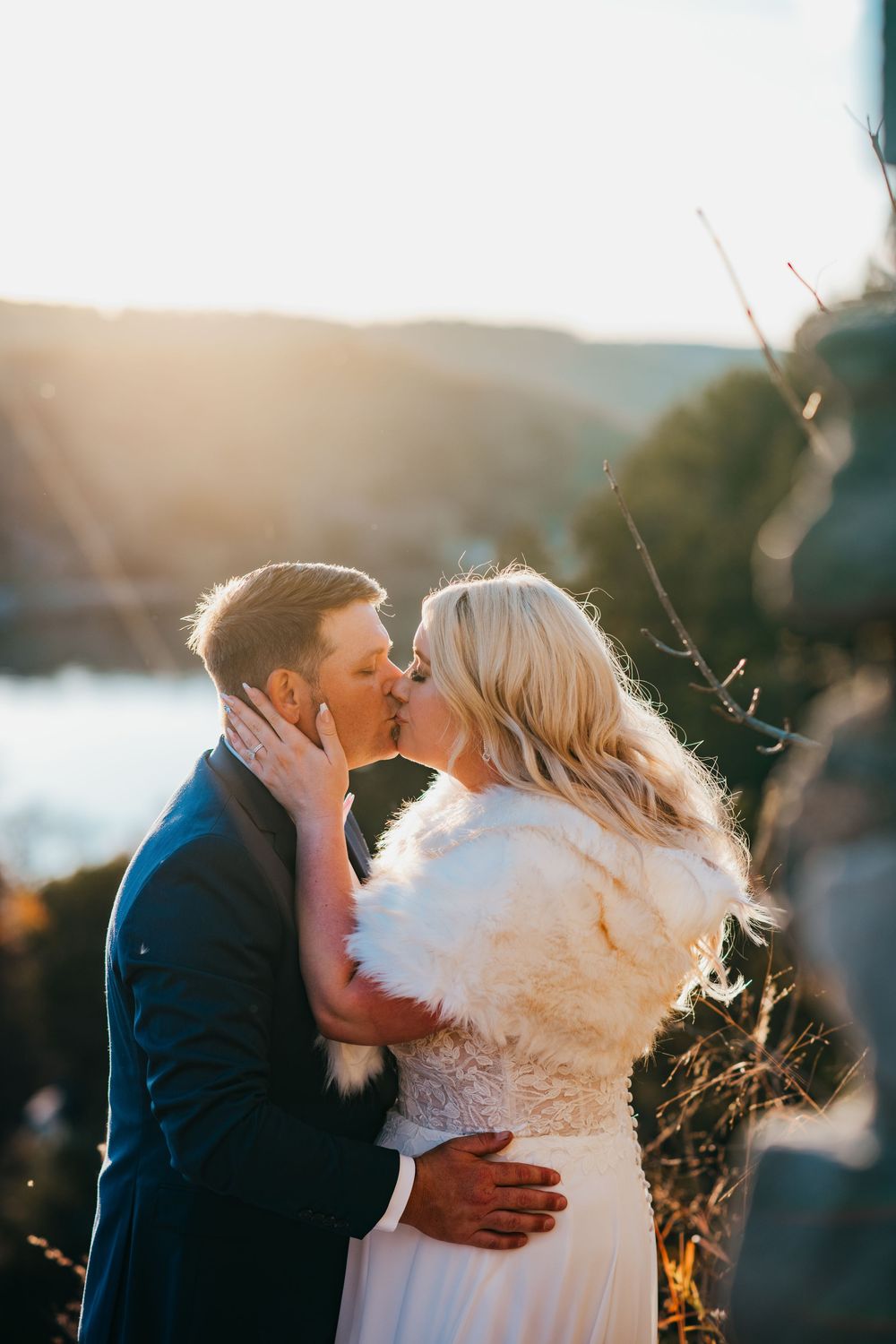 A romantic sunset kiss shared between a couple in wedding attire on a scenic hilltop overlook.