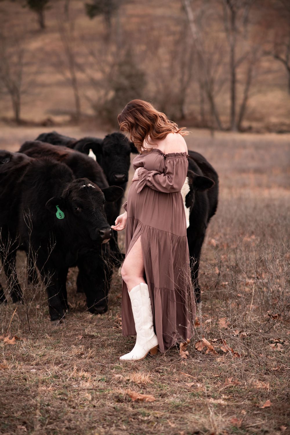 A person in a maxi dress and white boots stands in a field with black cattle against an autumn backdrop.