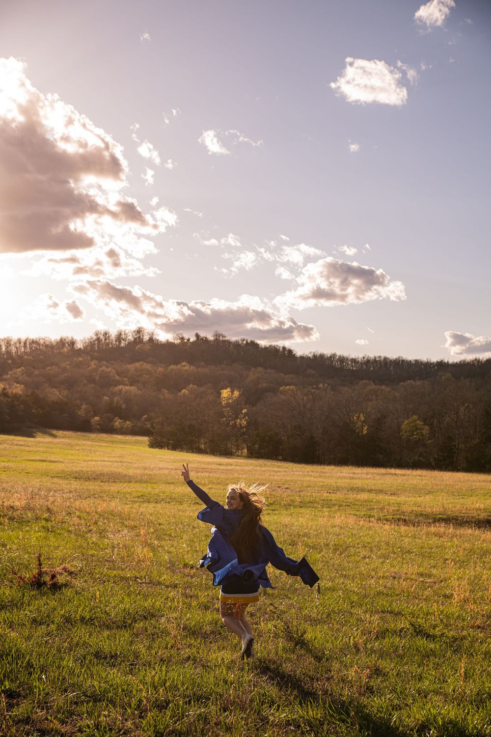 Person jumps joyfully in a sunny meadow at sunset with rolling hills and scattered clouds in the background.