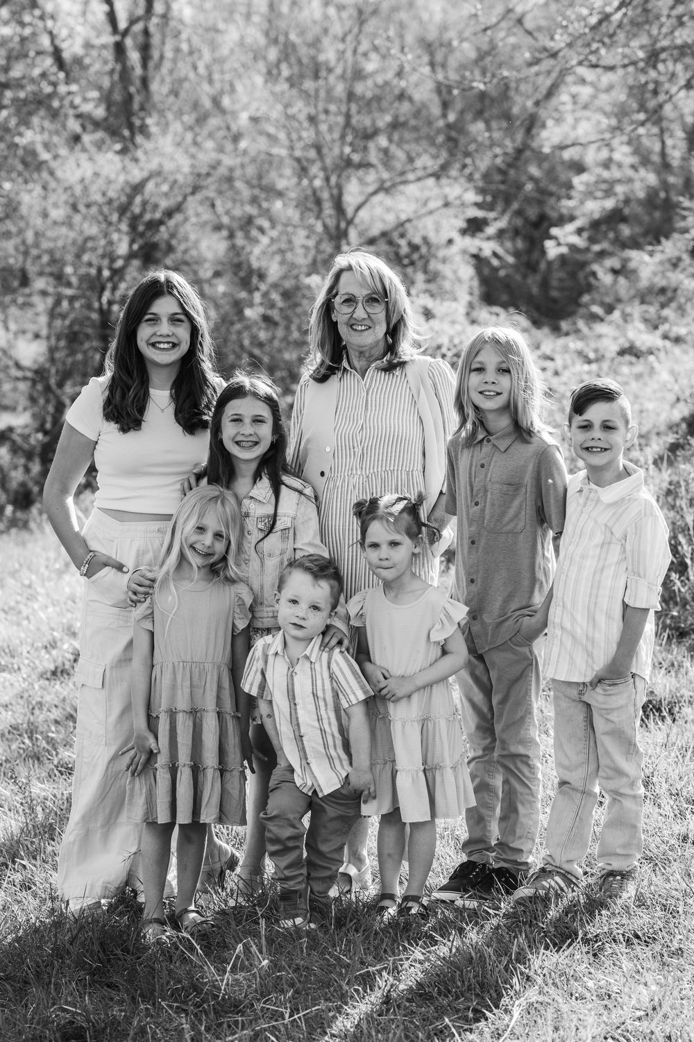 Black and white outdoor family portrait showing a group of seven people posed together in a natural wooded setting.