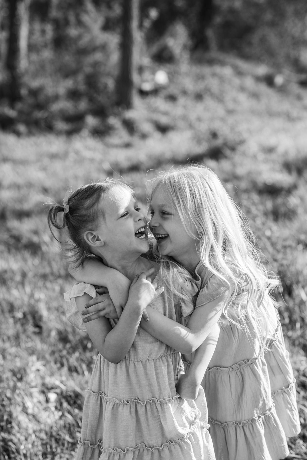 Two young children in matching ruffled dresses share a joyful laugh together outdoors in black and white photography.