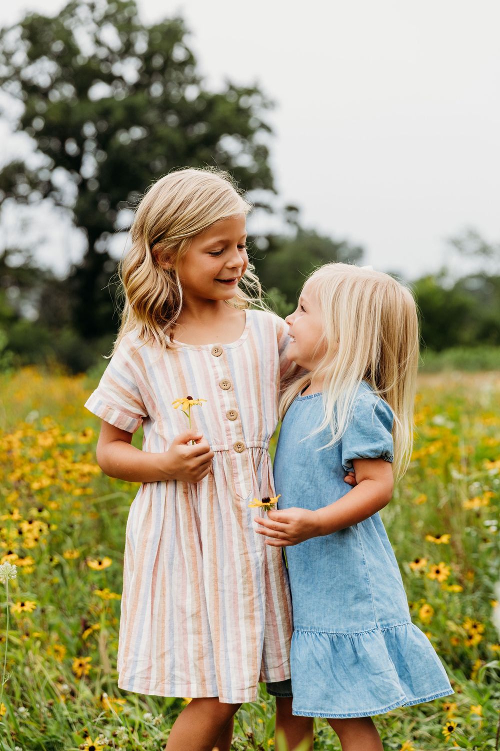 Two young children in summer dresses share a sweet moment among yellow wildflowers.