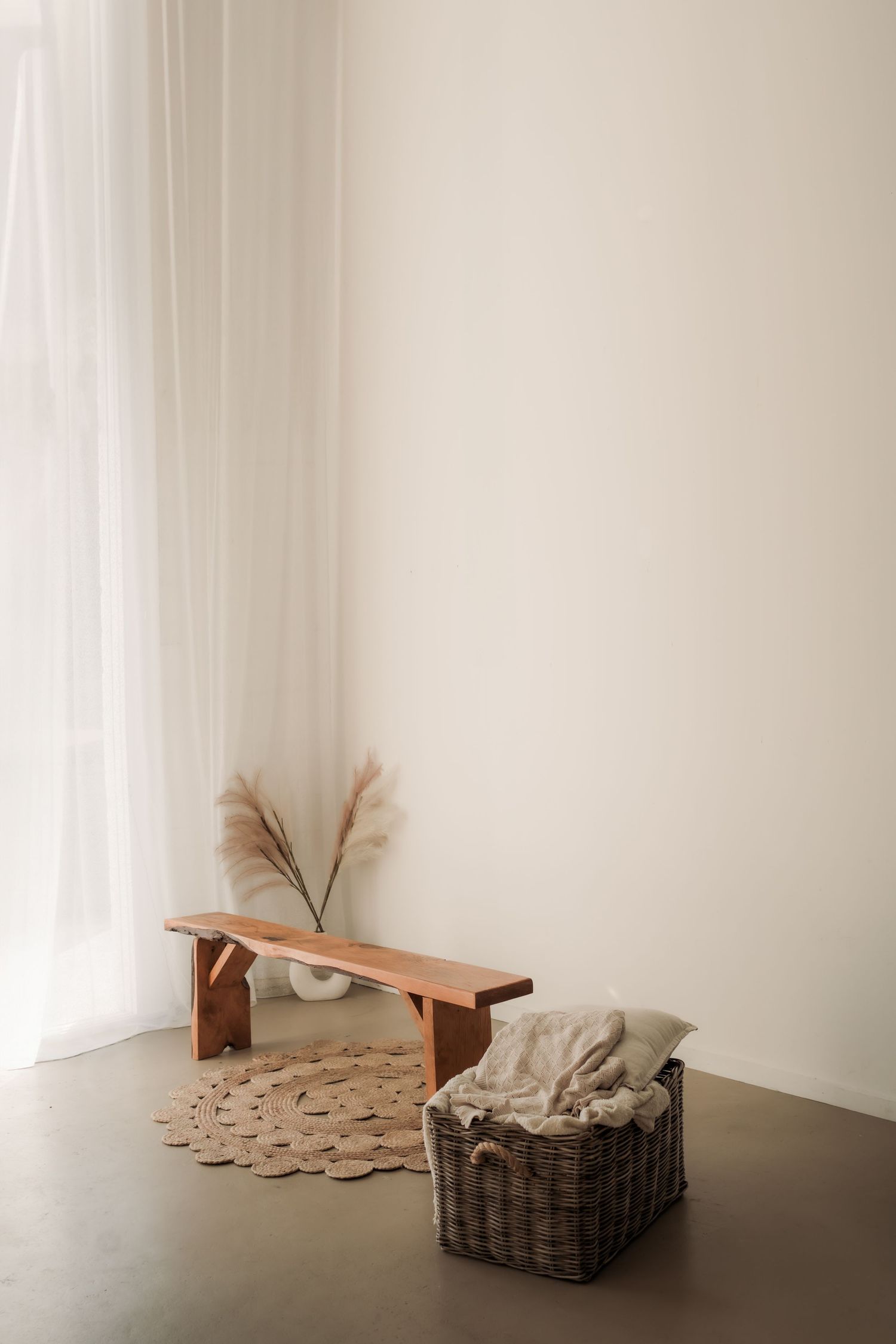 Minimalist interior scene with wooden bench, wicker basket, and dried pampas grass against white wall and natural light.