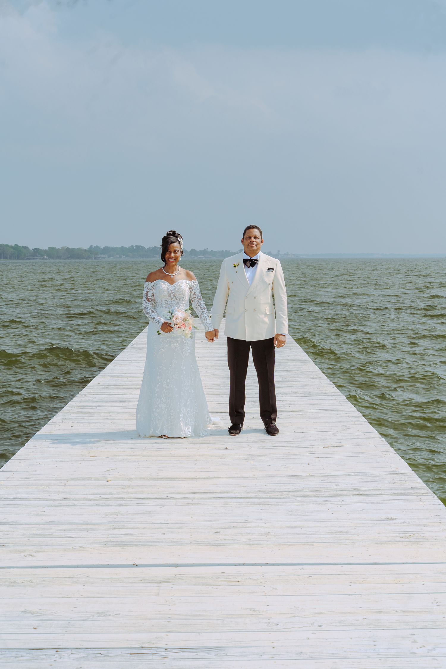 A couple in wedding attire stands at the end of a white wooden pier overlooking a vast body of water on their special day.